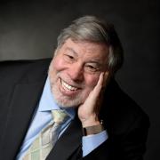 In a portrait with a dark studio background, Steve Wozniak leans forward, propping his face on his hand, smiling into the camera.